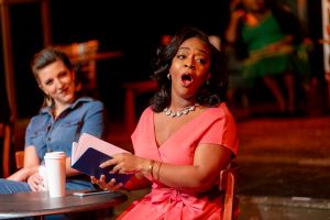 A woman in a pink dress sits at a table with an open book. She is singing to someone to her left. There is a white cup on the table and another woman sitting there in blue.