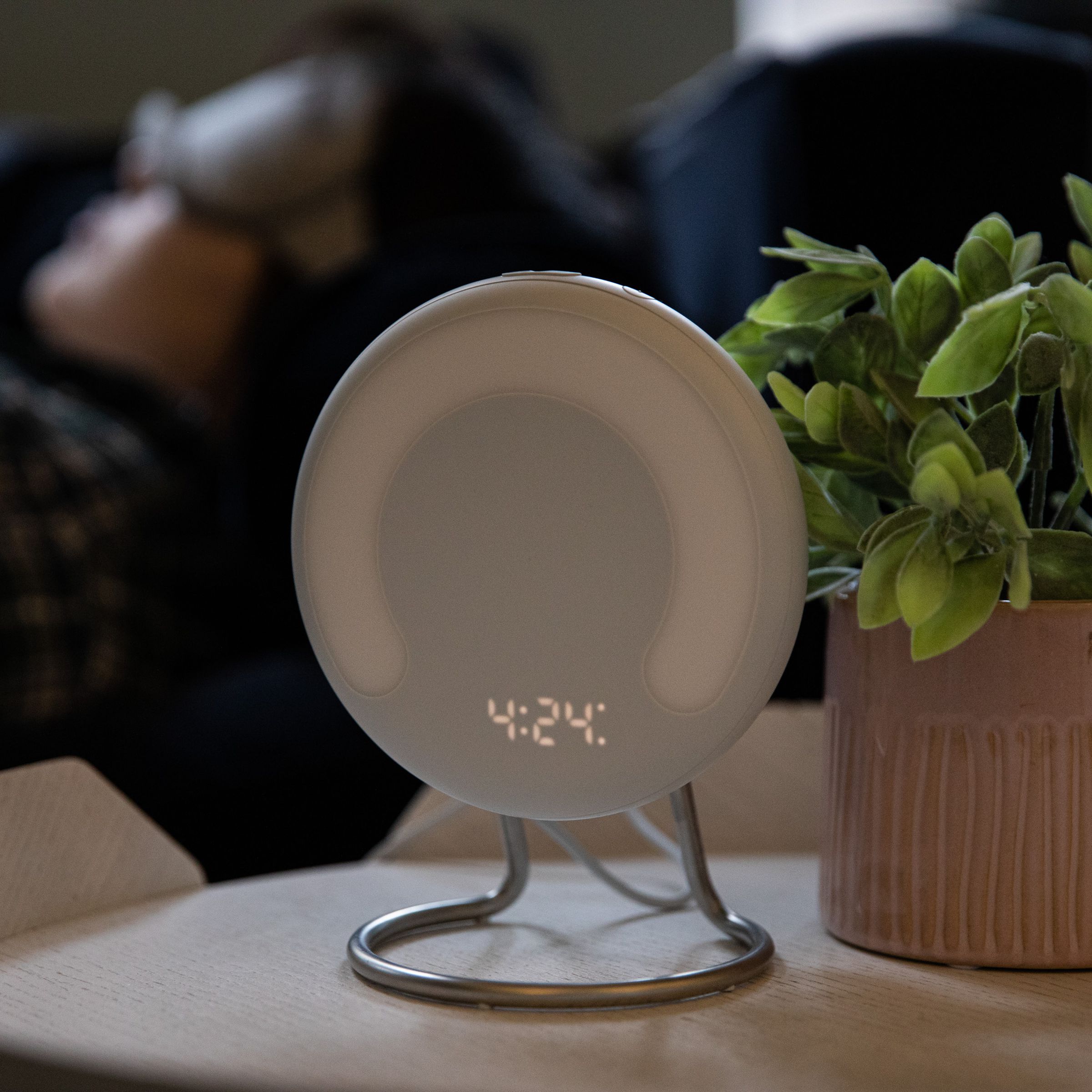 Amazon Halo Rise on a nightstand with a plant, while a person sleeps in the background.