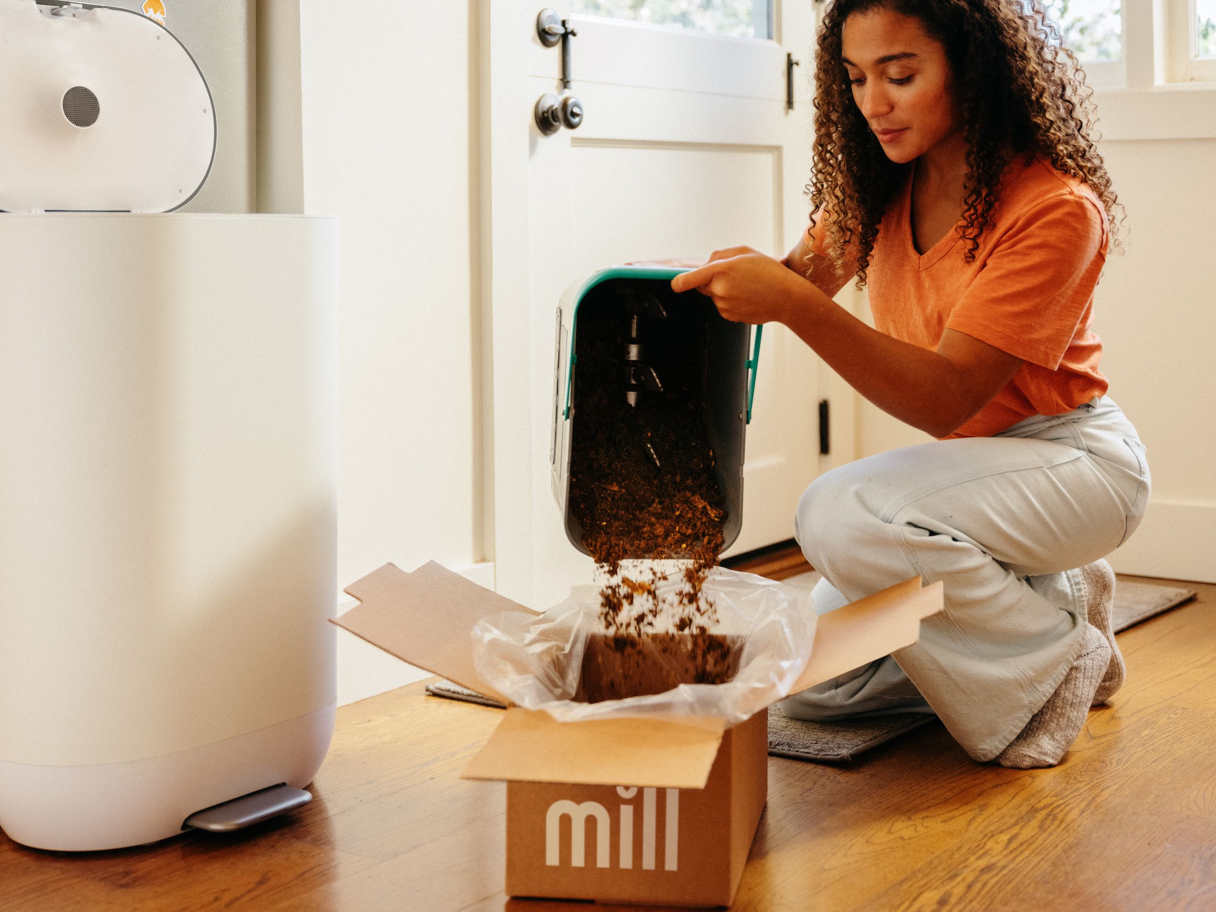 A woman kneeling pours food grounds out of a bin and into a cardboard box lined with a plastic bag.