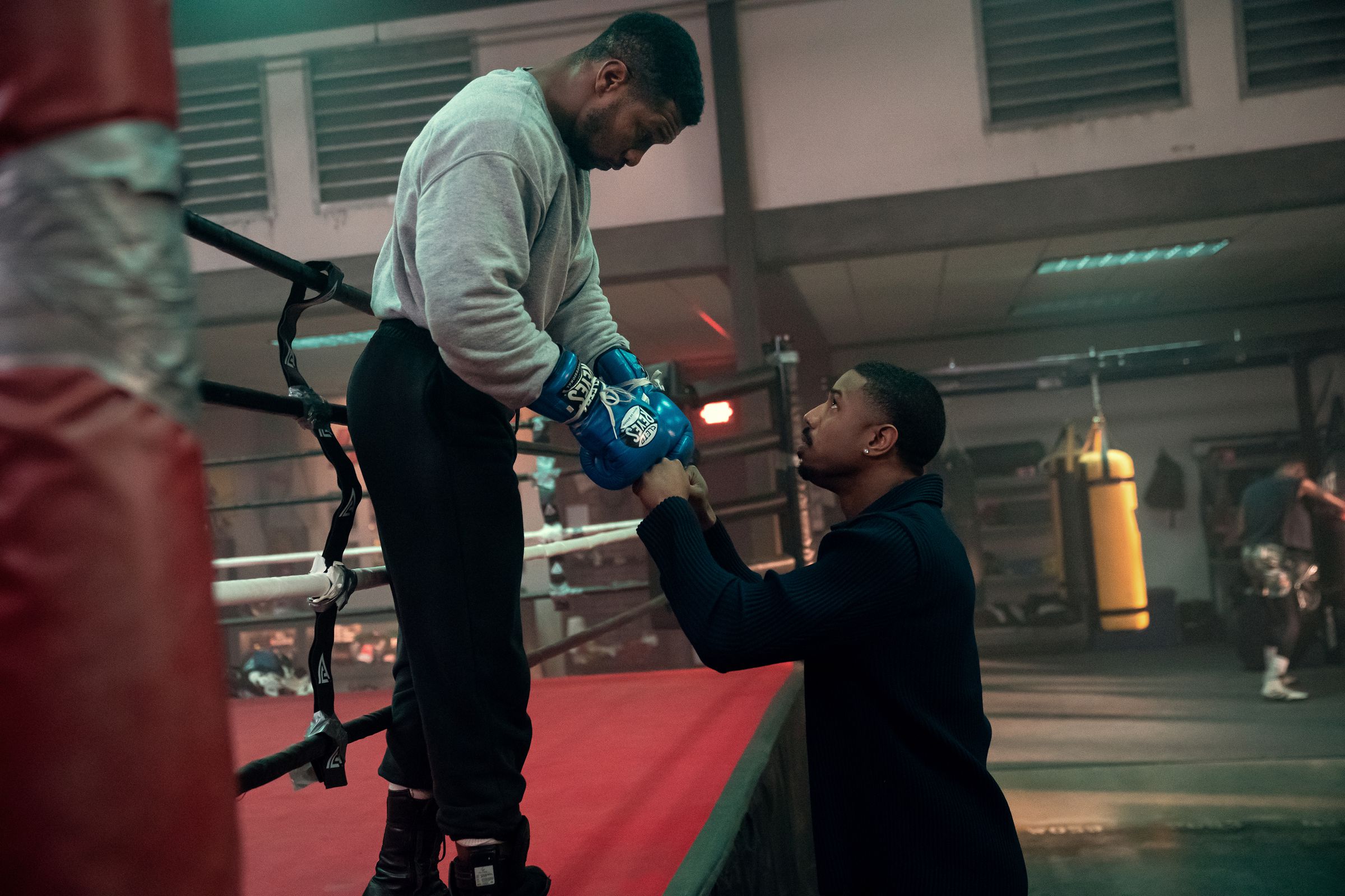 A man in a gray sweatshirt and black sweat pants leaning over a boxing ring to bump fists with a man wearing a black shirt.