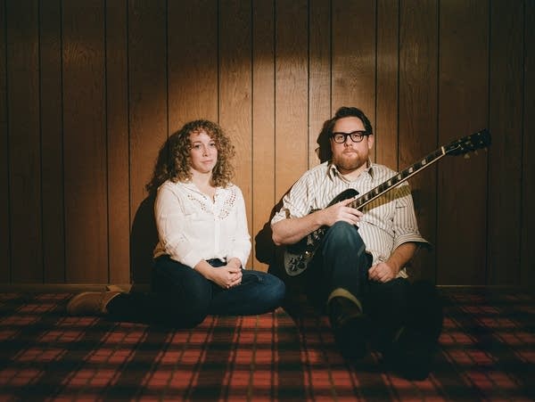 Two people sit spotlighted against a wood paneled wall on a checkered floor