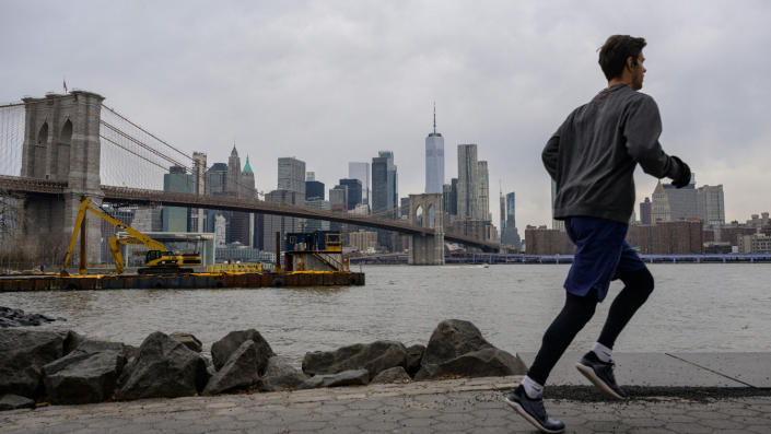 A man jogs in front of the skyline of lower Manhattan and the Brooklyn Bridge.