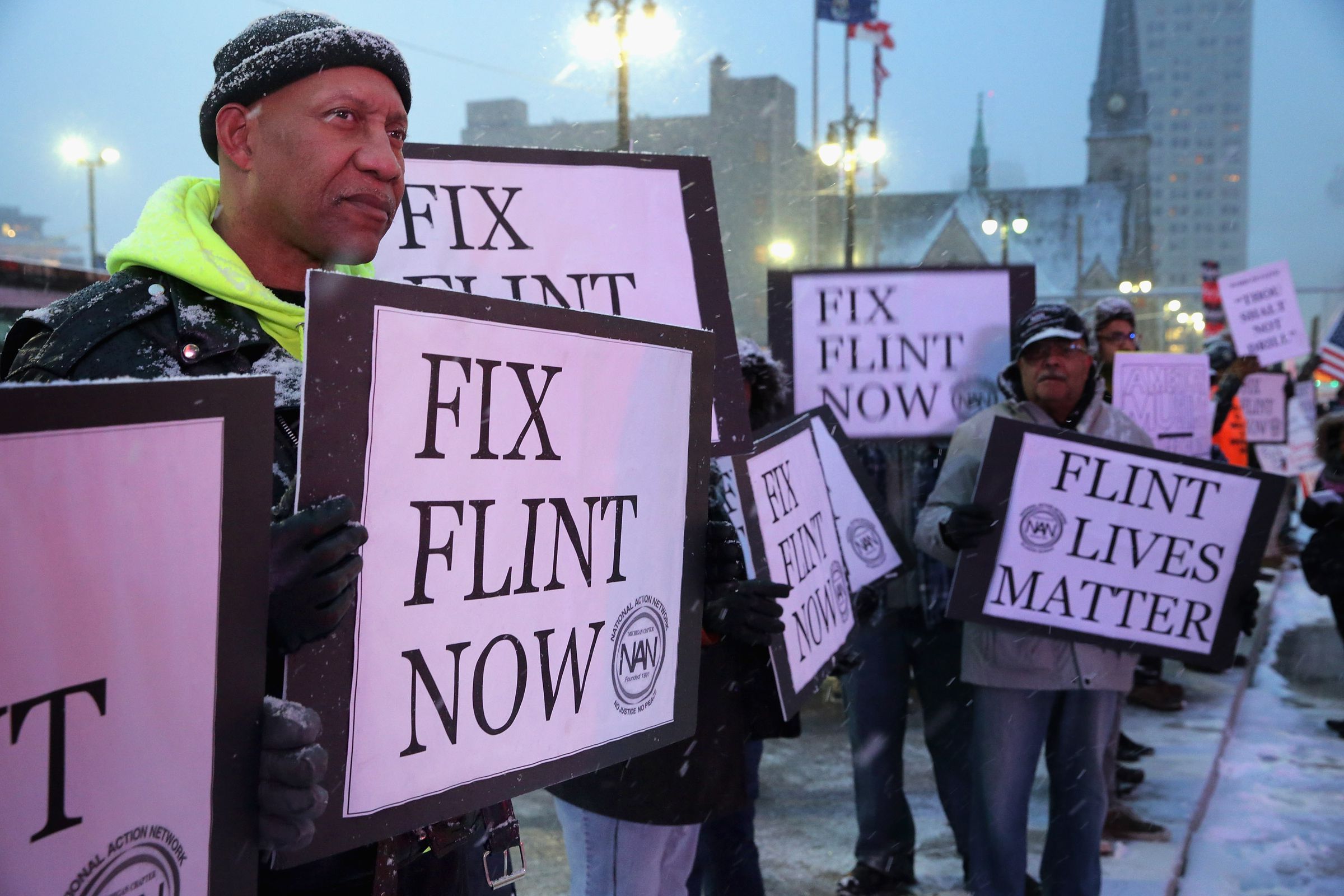 People stand side by side carrying signs that say “Fix Flint Now” and “Flint Lives Matter”