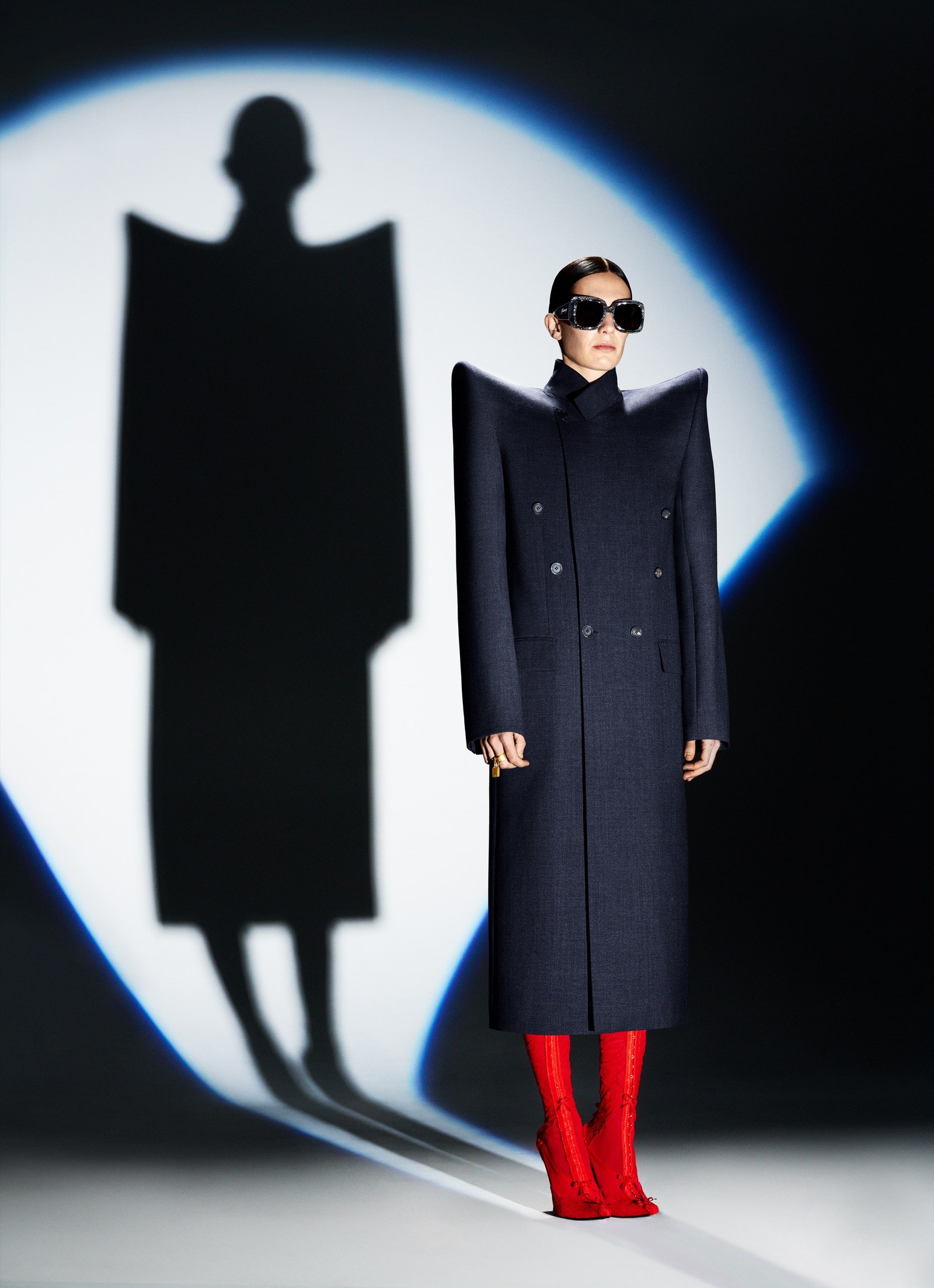 A model stands in a spotlight wearing a coat with high shoulders.