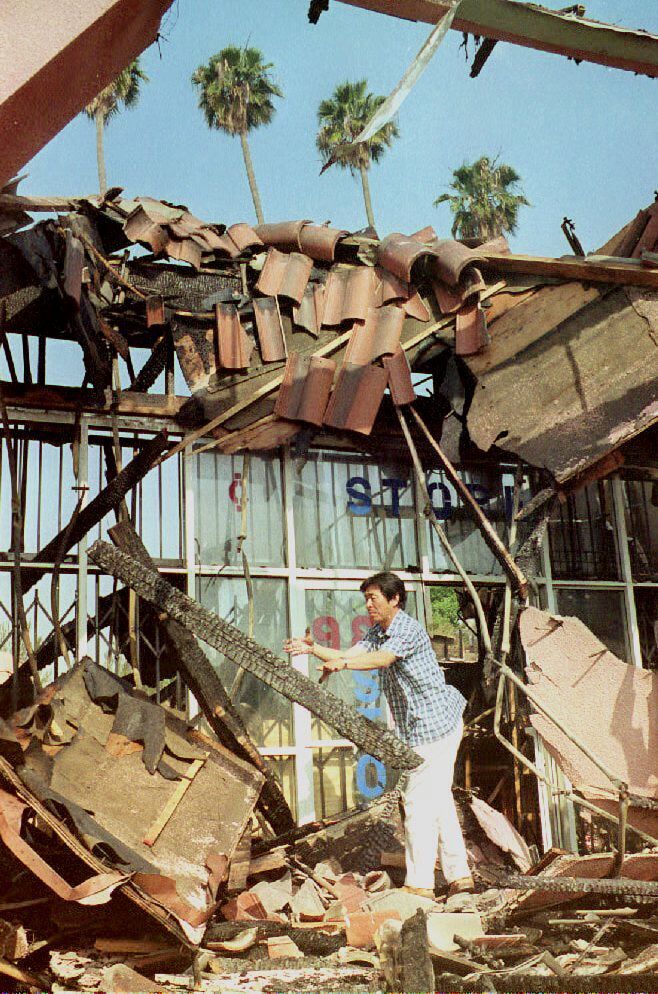 A Spanish-tyle roof is in shatters amid debris from a storefront as an Asian man tries to clean up from the destruction.