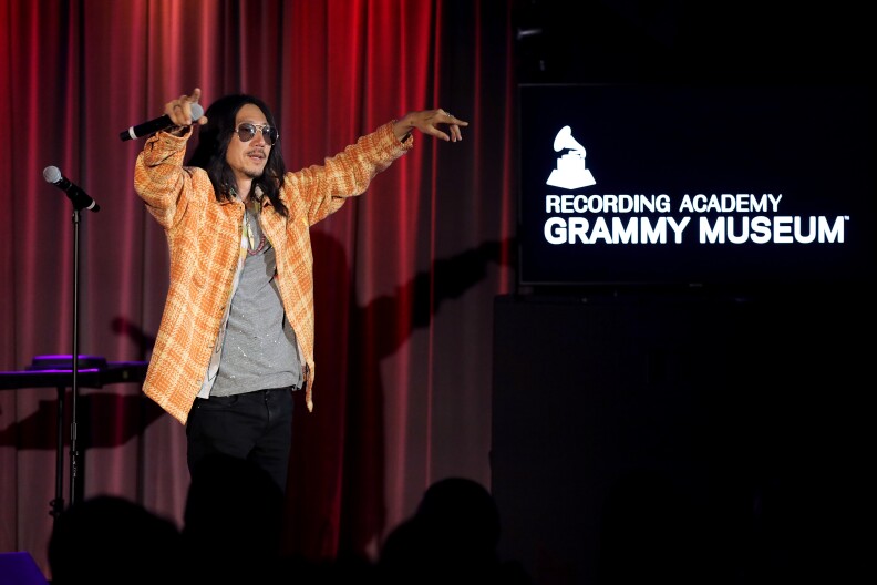 An Asian man has sunglasses and facial hair. He wears sunglasses and and Orange jacket over a T-shirt. A sign for the Recording Academy Grammy Museum is visible to the right.