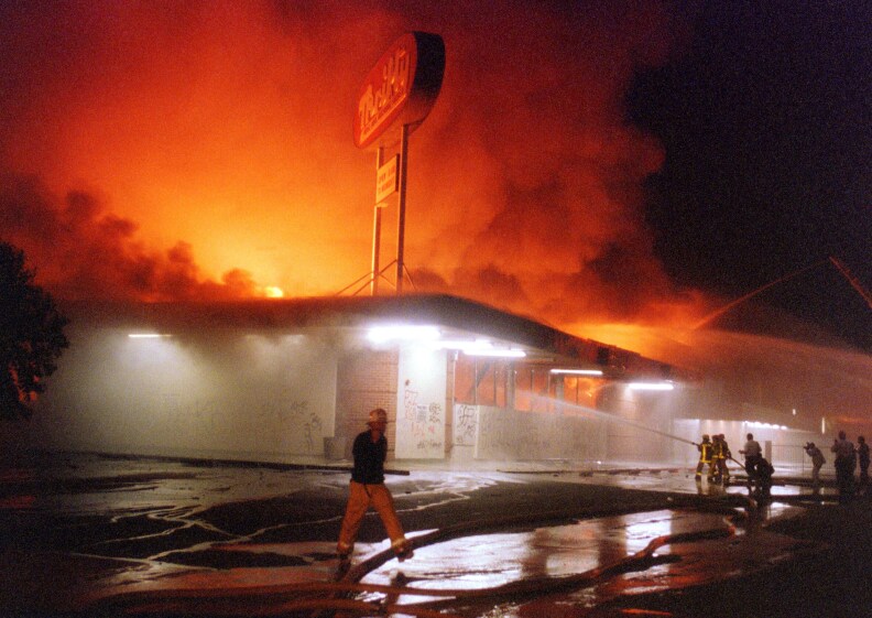 Flames roar from a Thrifty Drug store as firefighters point a hose toward the fire.