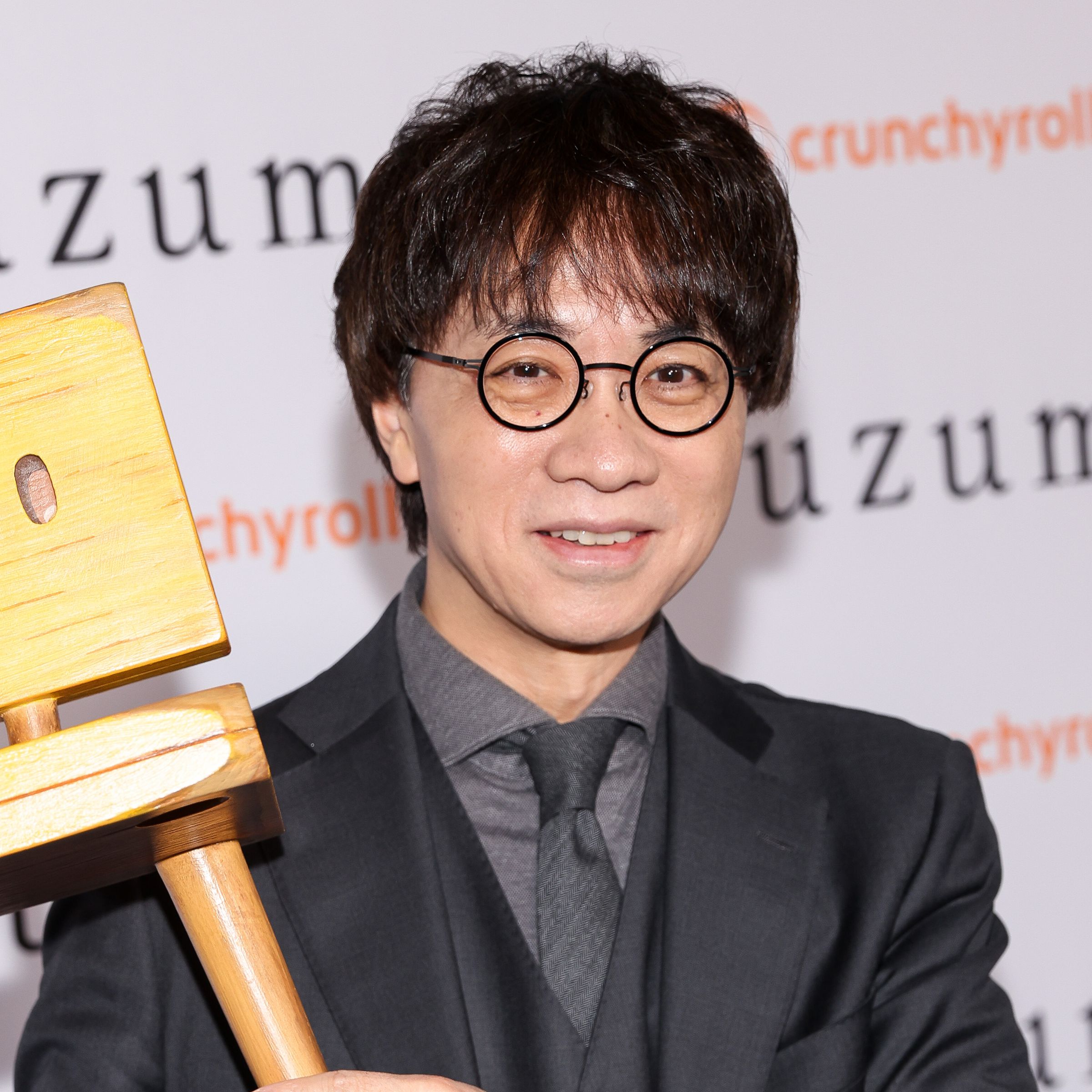 A bespectacled man in a black suit with a steel gray shirt and tie holding up a wooden chair.