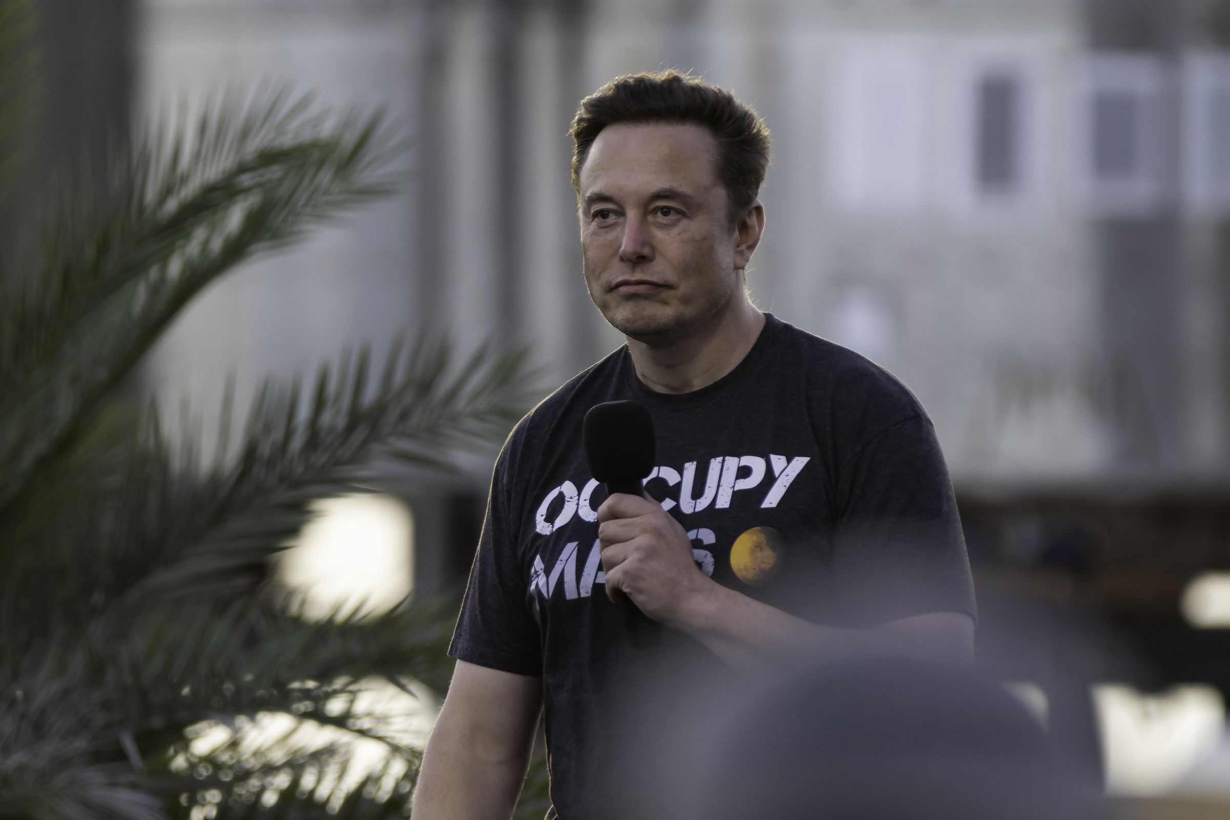 Elon Musk holds a microphone to his face while wearing a T-shirt that says “Occupy Mars.”