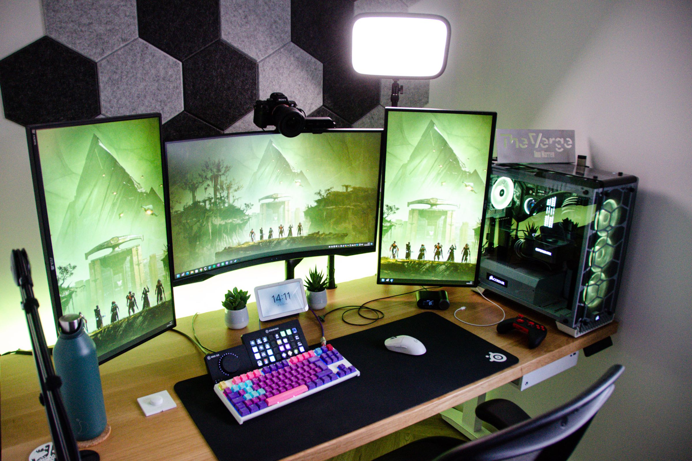 A desk from an overhead view with three monitors, a colorful keyboard in the center, a PC on the right, and various other tech.