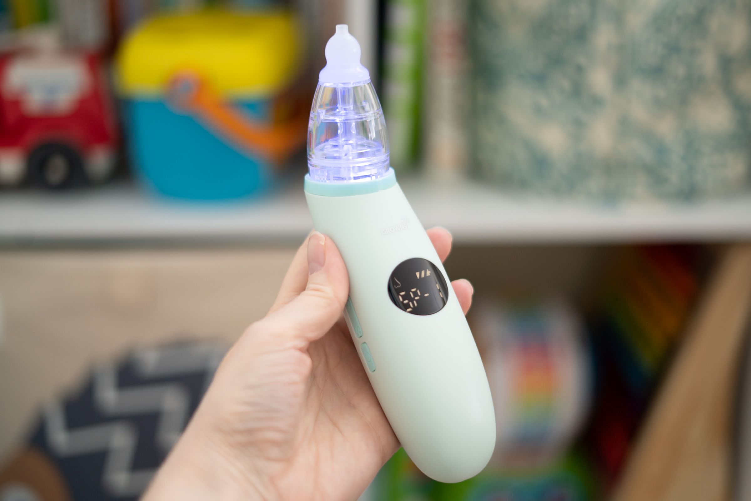 Hand holding a nasal aspirator in front of a colorful background of childrens’ toys and books.