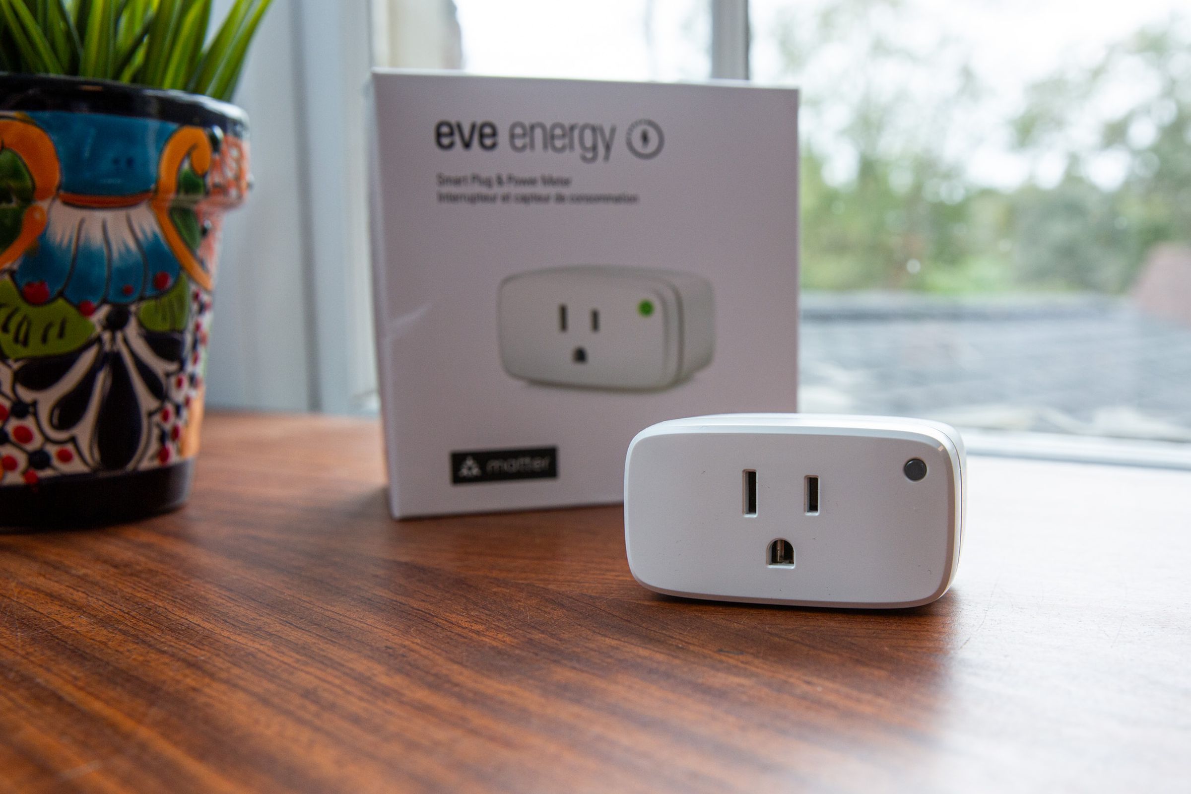 A white smart plug in front of its box on a desk next to a window.