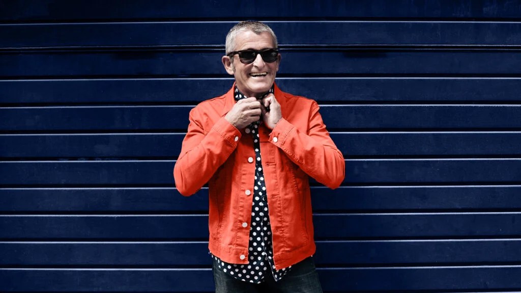 happy older man wearing sunglasses adjusting black and white polka dot shirt under orange jacket