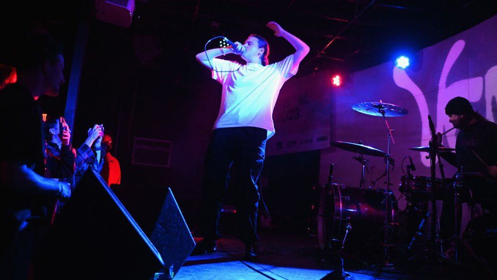 Venbee performing on-stage. She's wearing a white t-shirt and black trousers. She's bathed in a purple light, her eyes are closed and she has one arm, bent above her head while the other hand holds the microphone close to her mouth.