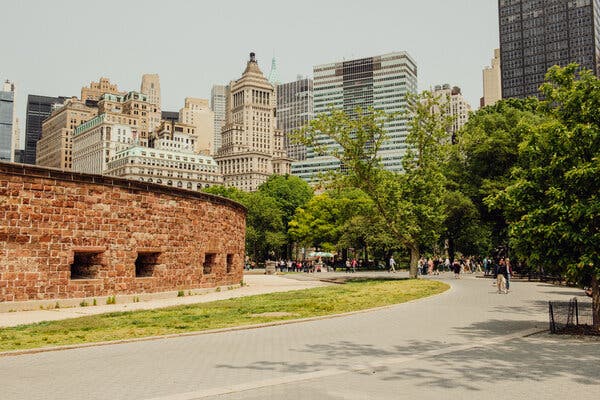 A view of an urban park with a wide sidewalk and a centuries-old, low, brick, circular construction with square openings along the bottom. The sidewalk is surrounded by grass and trees, and on a bright day, a dense urban cluster of buildings rises in the distance.