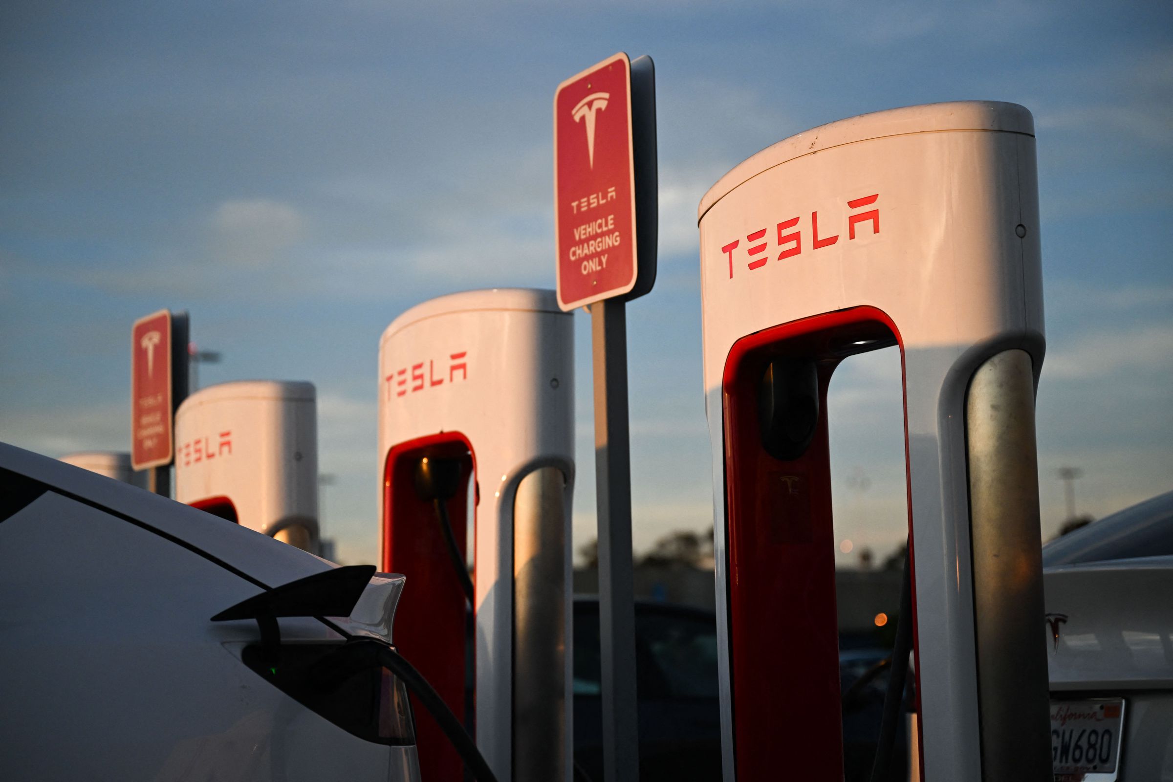 Tesla Supercharger station against a sunset and blue sky, and a Model Y is charging at one of the three stations in the shot