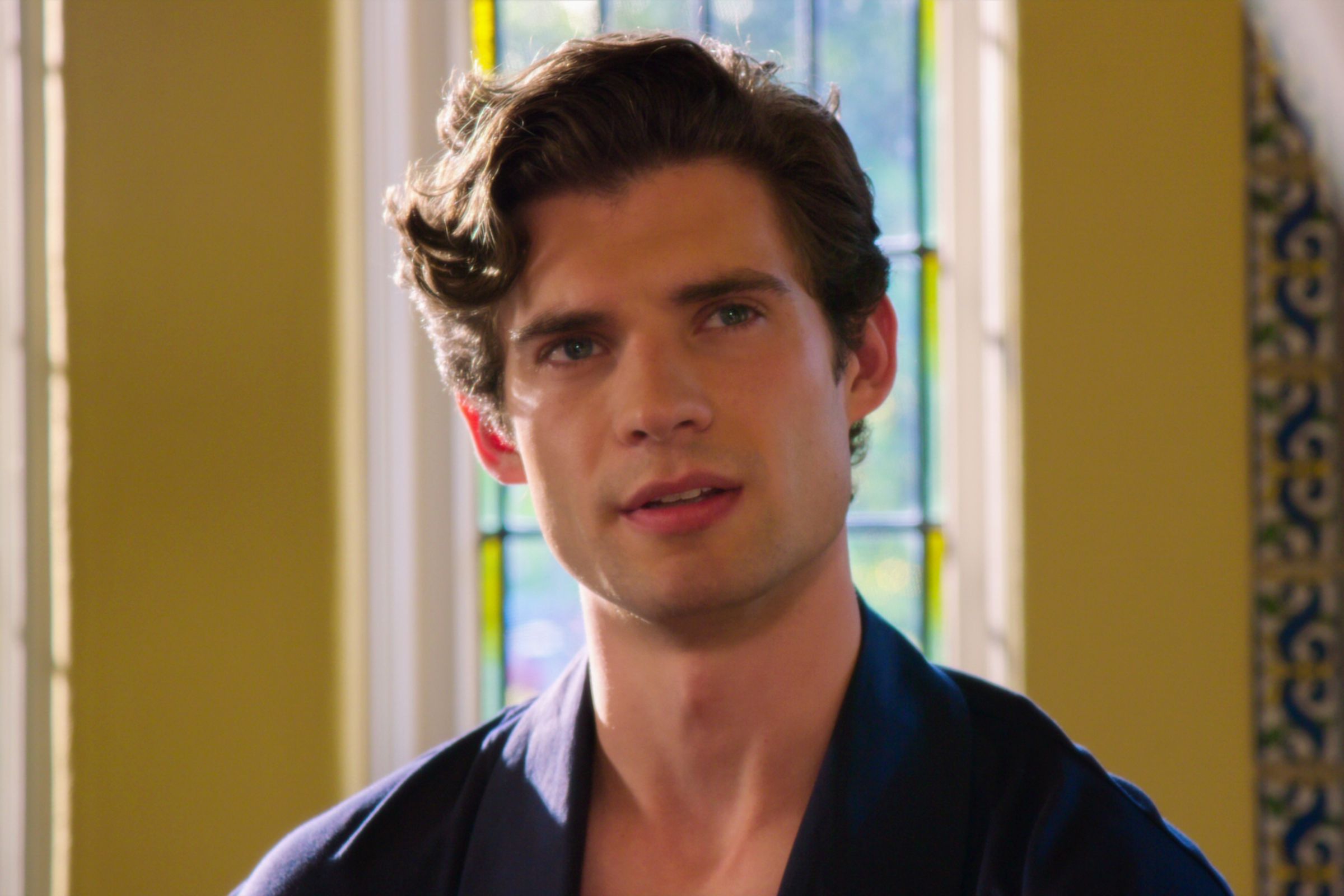 A man wearing a blue shirt sitting in front of a set of windows through which sunlight is shining.