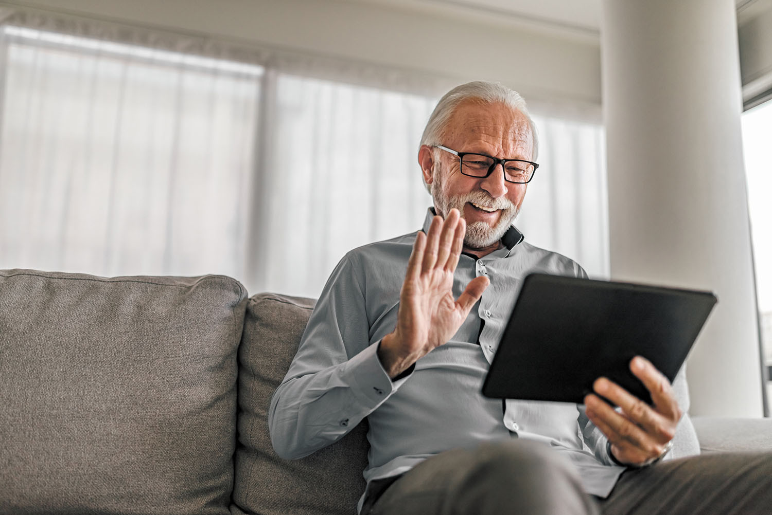 photo of a senior man at home using a tablet for a video call, smiling and waving