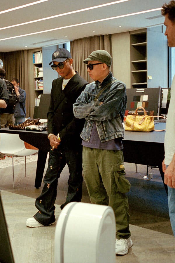 Mr. Williams and Nigo appear to be intently studying something in the workshop. Mr. Williams is wearing a black jacket and pants; Nigo wears a denim jean jacket and army green cargo pants. Both have on ball caps and sunglasses. 