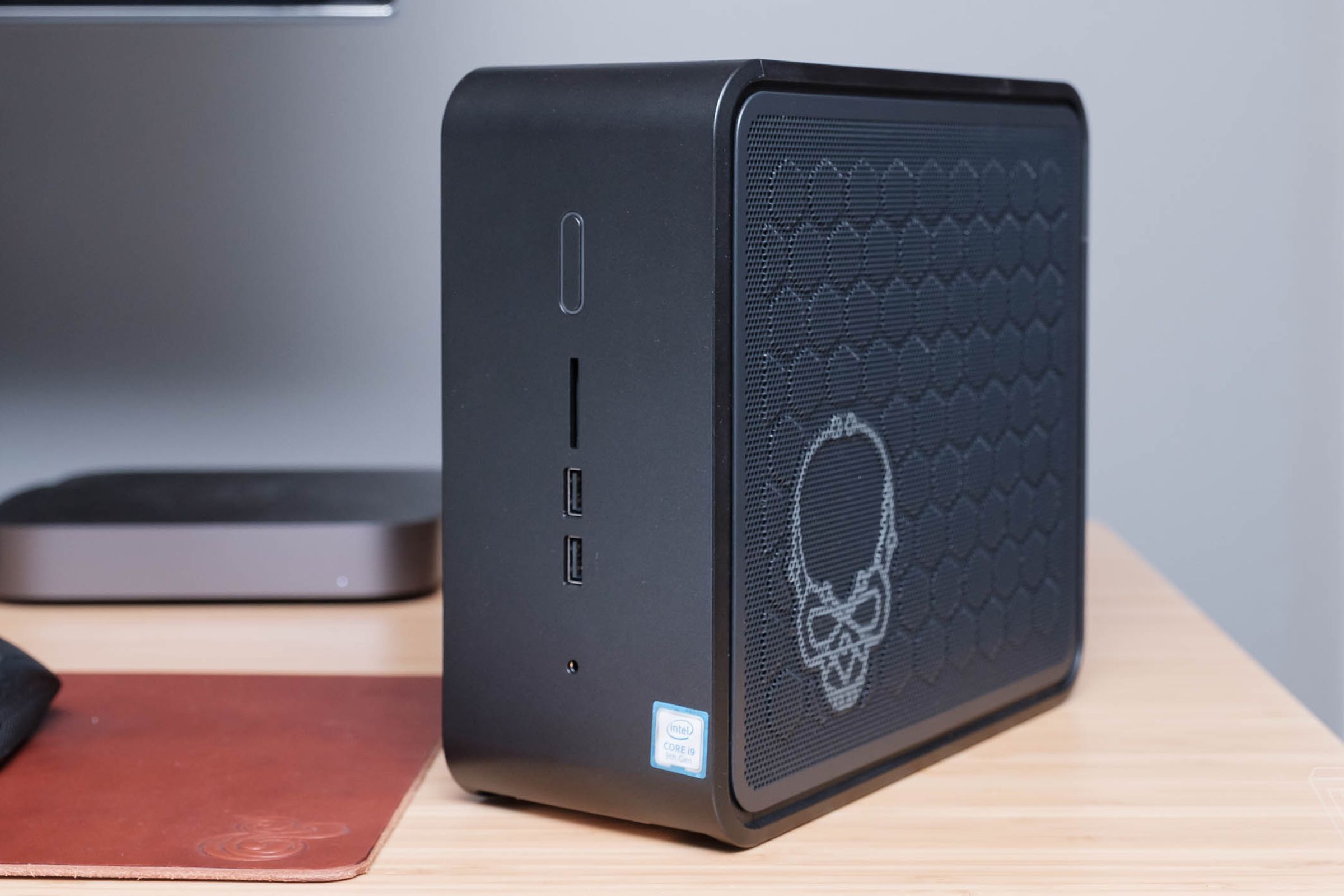 Intel NUC 9 computer standing vertically on a wood desk with a skull etched on the side and a gray Mac Mini in the background