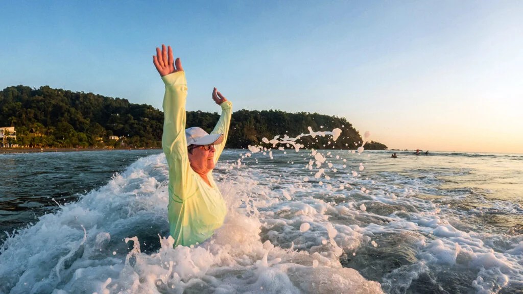 An older person throws both arms in the air in joy while standing in the ocean water on a sunny day