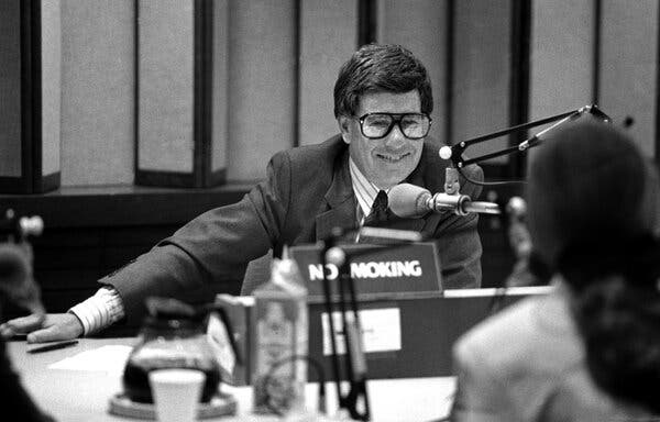 A black-and-white photo of Robert Sherman smiling as he sits across a table from a young woman with her back to the camera. A mic hangs in front of Mr. Sherman.