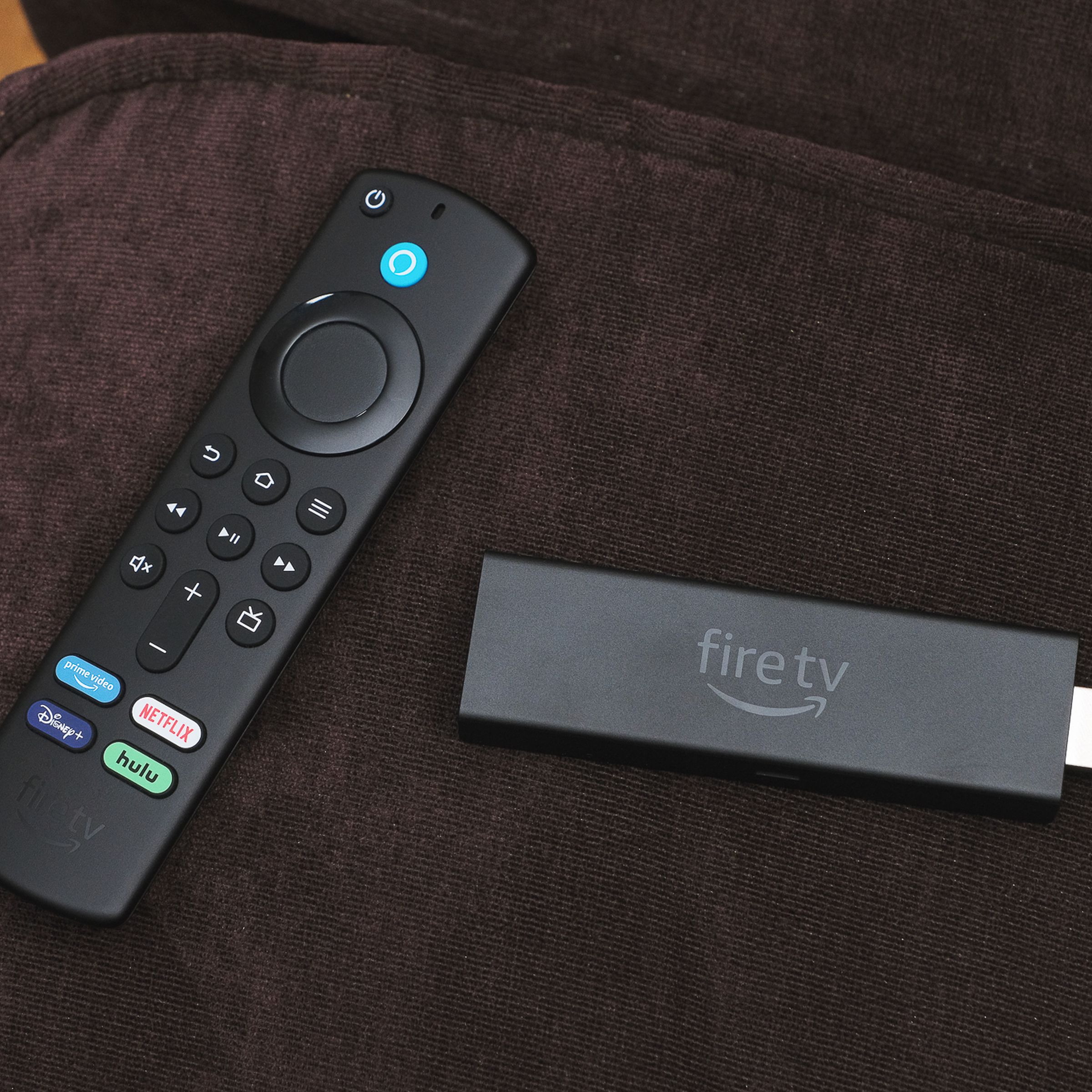 A close-up of the Amazon Fire TV Stick 4K Max streaming dongle beside its remote control on a brown couch.