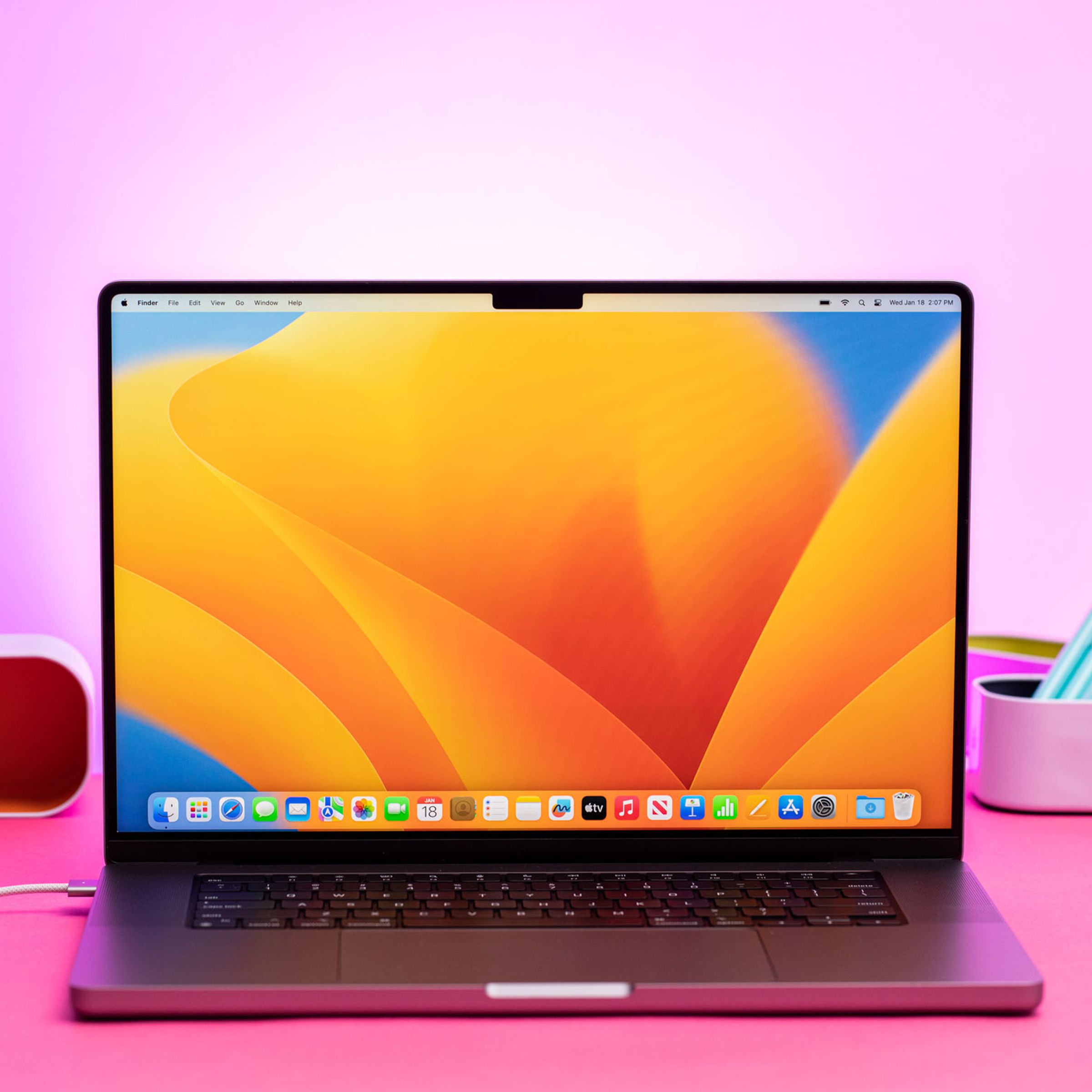 The MacBook Pro 16 (2023) on a pink table. The screen displays a blue and yellow desktop pattern.