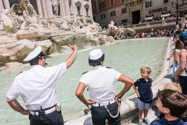 A police officer in a white uniform blows a whistle and waves a finger at visitors packed up against the edge of the basin of a large white stone fountain filled with bluish water.