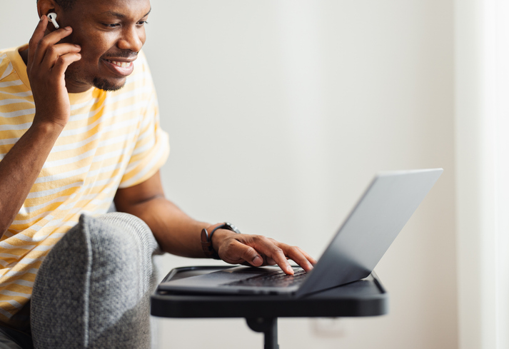 Smiling African American Man Putting Earbuds in His Ear and Using His Laptop Computer at Home