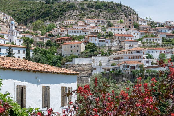 A village of white-walled buildings with red-tile roofs and wooden shutters extends up a rocky hill covered with scrubby bushes and trees. In the foreground, there are bright red flowers, and about halfway up the hill there is a skinny white tower with a light blue dome on top.