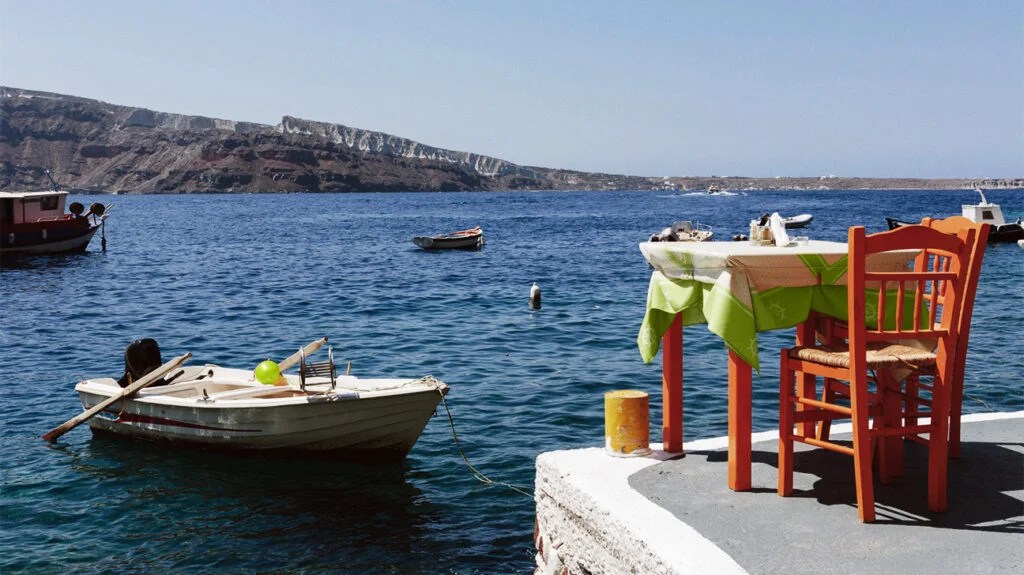 Mediterranean landscape with boats in the harbour and a small table and chair on the dock looking out to sea