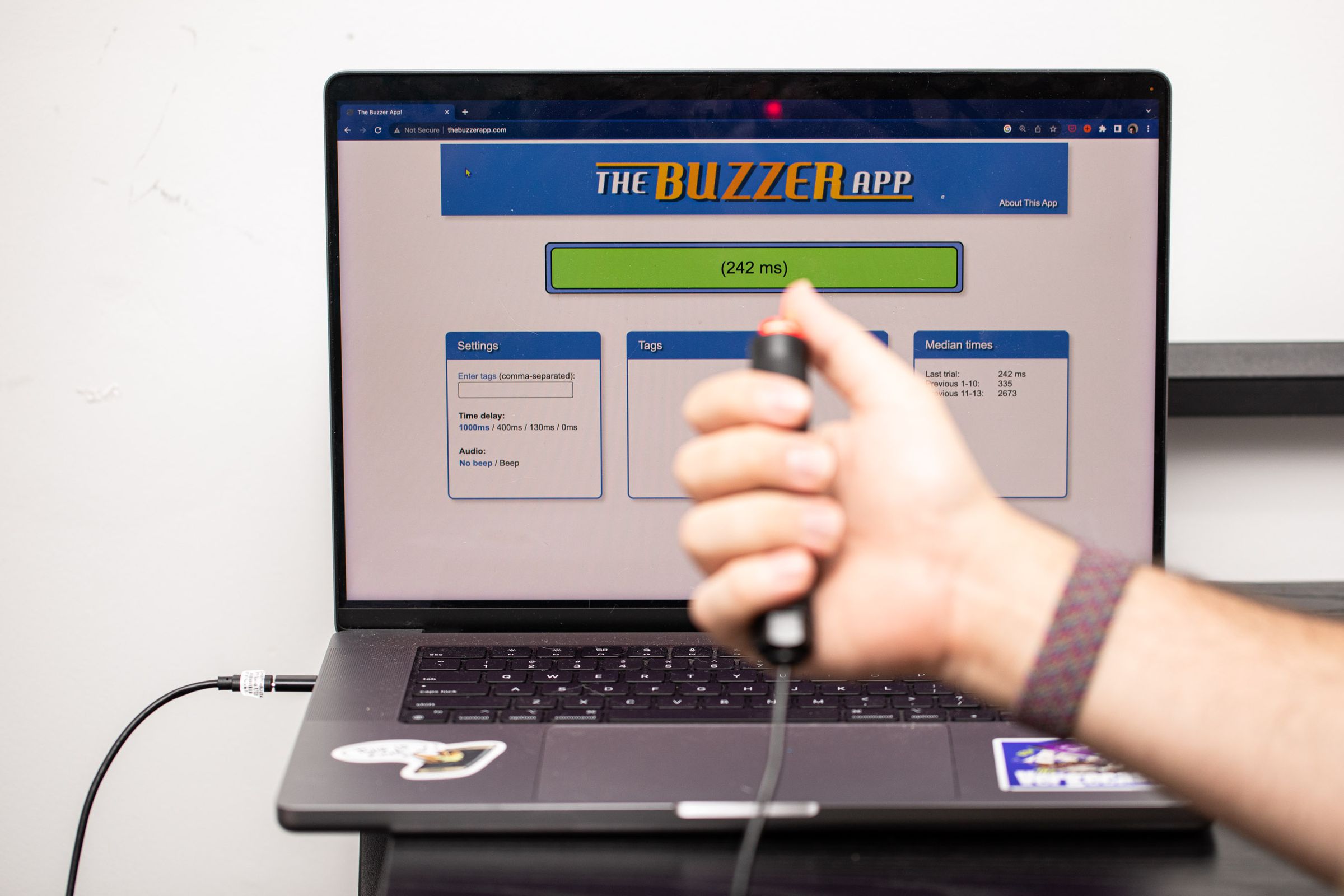 A laptop with the Buzzer App open in a browser with a hand holding the Delcom buzzer. The score is 242 milliseconds.