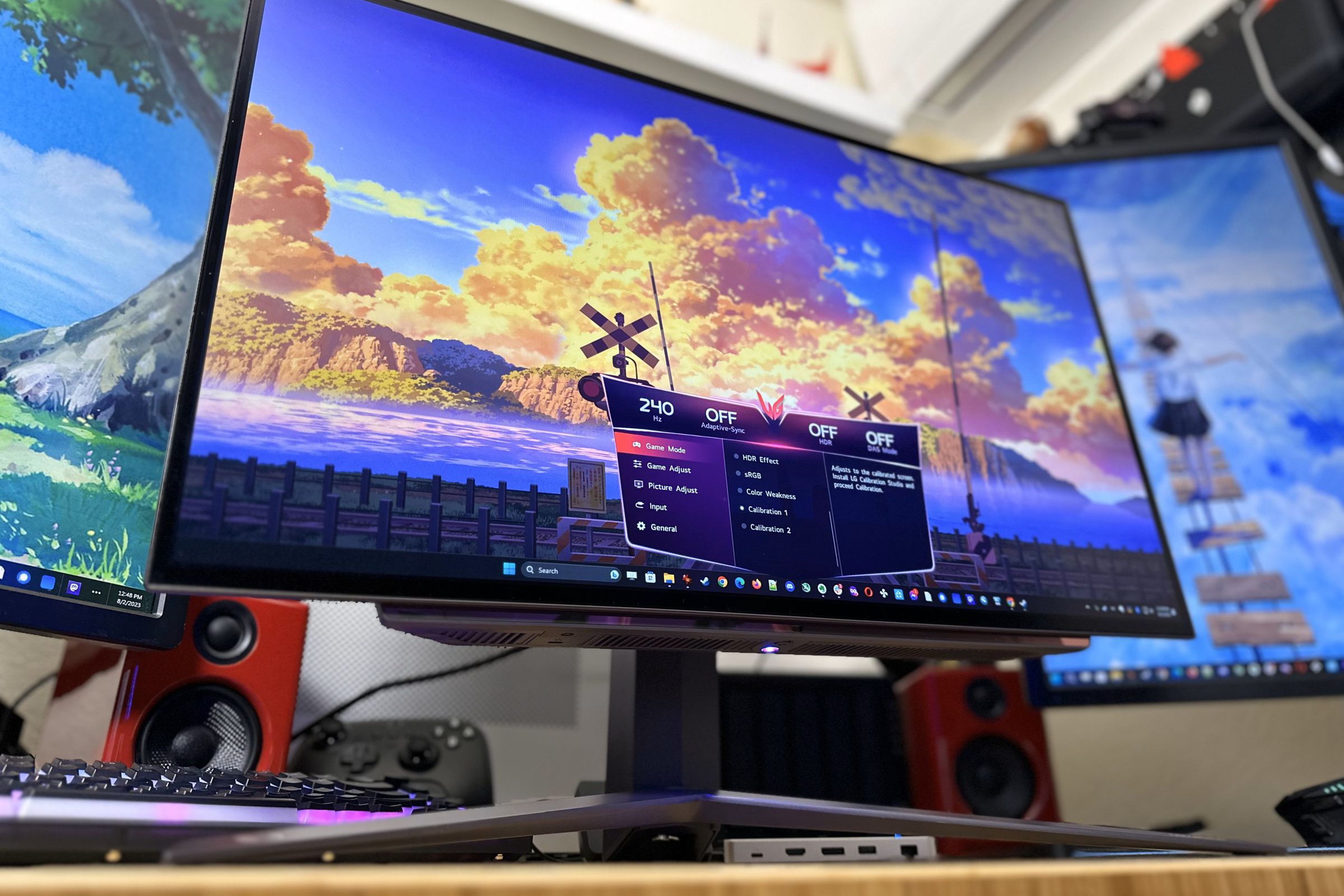 A bright, colorful OLED monitor with a big V-shaped stand underneath, flanked by two other monitors in portrait mode. The main monitor has a colorful sky on screen with big orange sunset-lit clouds and an on-screen display showing it’s running at 240Hz.