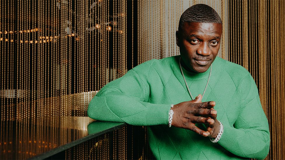 Akon wearing a green top with his hands joined together at the fingertrips. He is wearing a gold necklace as he looks into the camera. The background is shiny gold pearls dropping from top to bottom, as his right elbow is perched on a table.