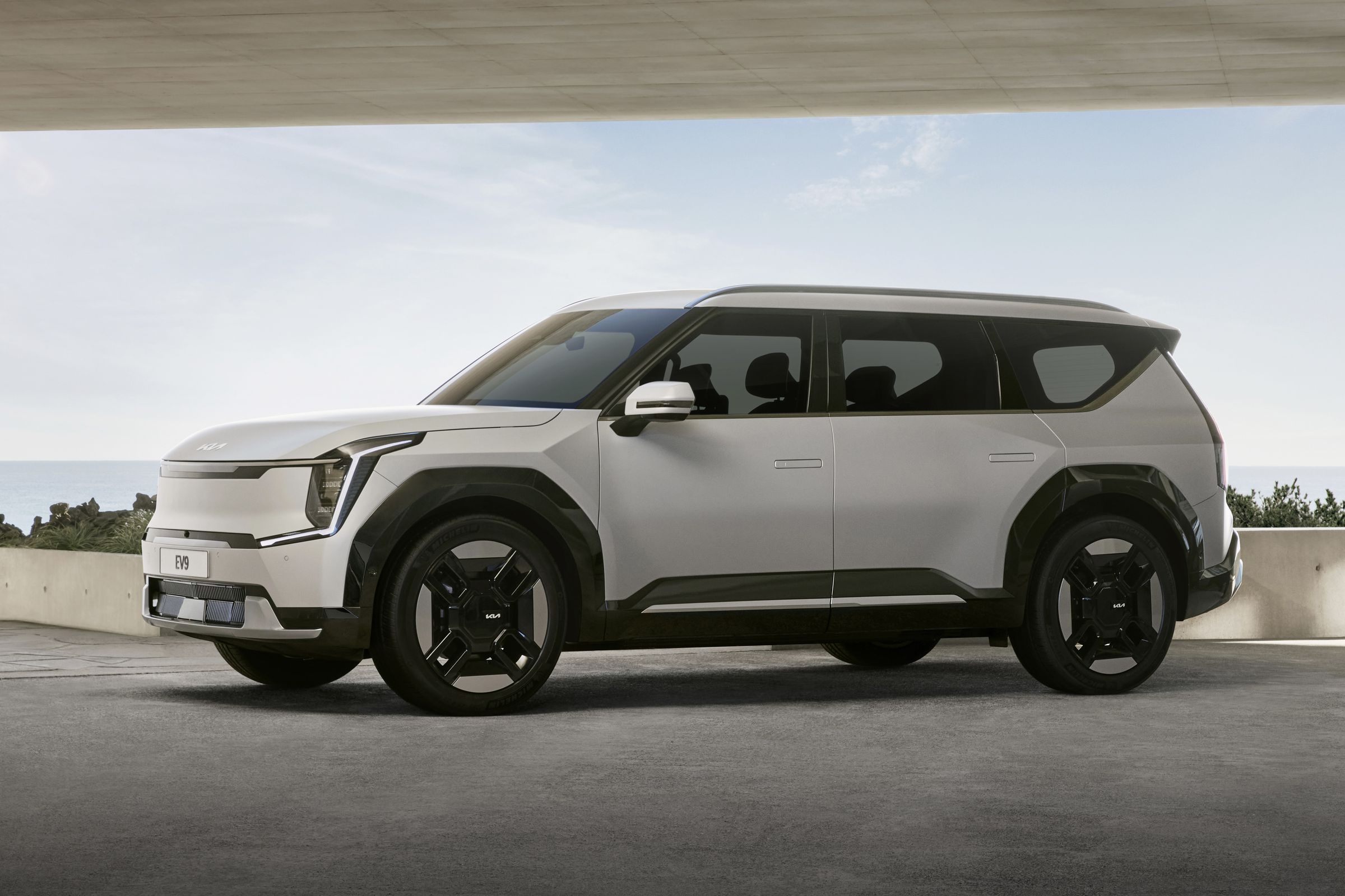 white kia ev9 suv on a concrete pad overlooking the sky. car has flush door handles, a paint-colored smooth grille, big rims with a flat aerodynamic finish, and looks really big.