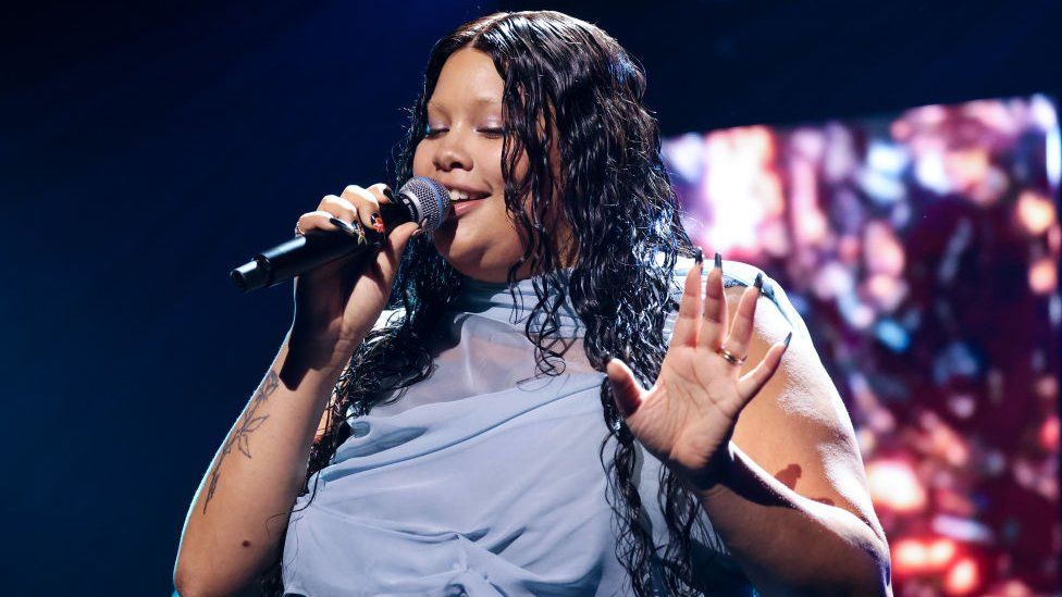 Shygirl performing at the Mercury Prize. Real name Blane Muise, Blane wears a pale blue chiffon dress which drapes around her neck. She has long, dark wavy hair which she wears loose over her shoulders. She holds a microphone to her face with her right hand, revealing a rose forearm tattoo, and holds her left palm up. Dark staging can be seen behind her and she is lit by a spotlight.