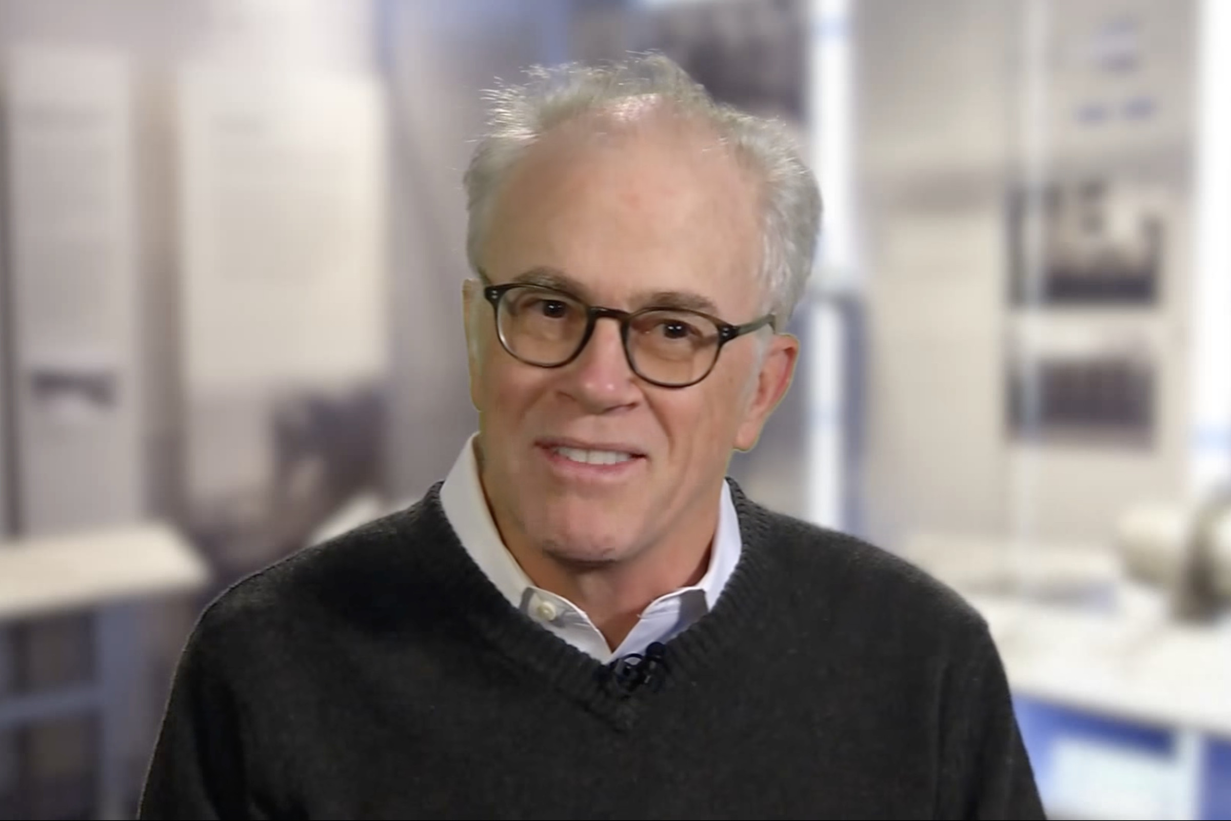 A screenshot of Dennis Austin smiling. His gray hair is close-cropped and quite thin on top, and he is wearing dark glasses a dark sweater over a dress shirt.