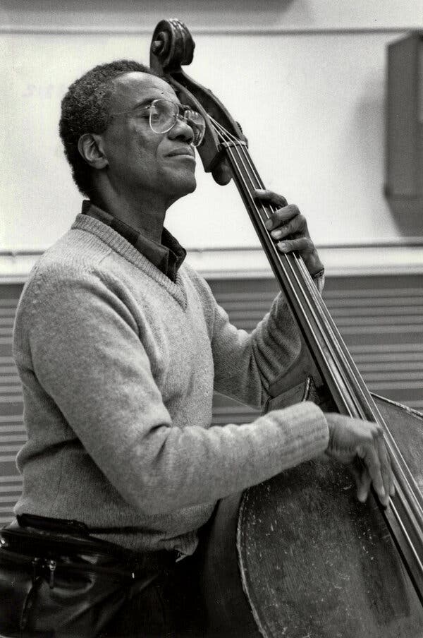 A black-and-white photograph of Richard Davis playing a stand-up bass. A tall man with flecks of gray in his hair, he is wearing a light-colored sweater and large glasses and has a serious look on his face.