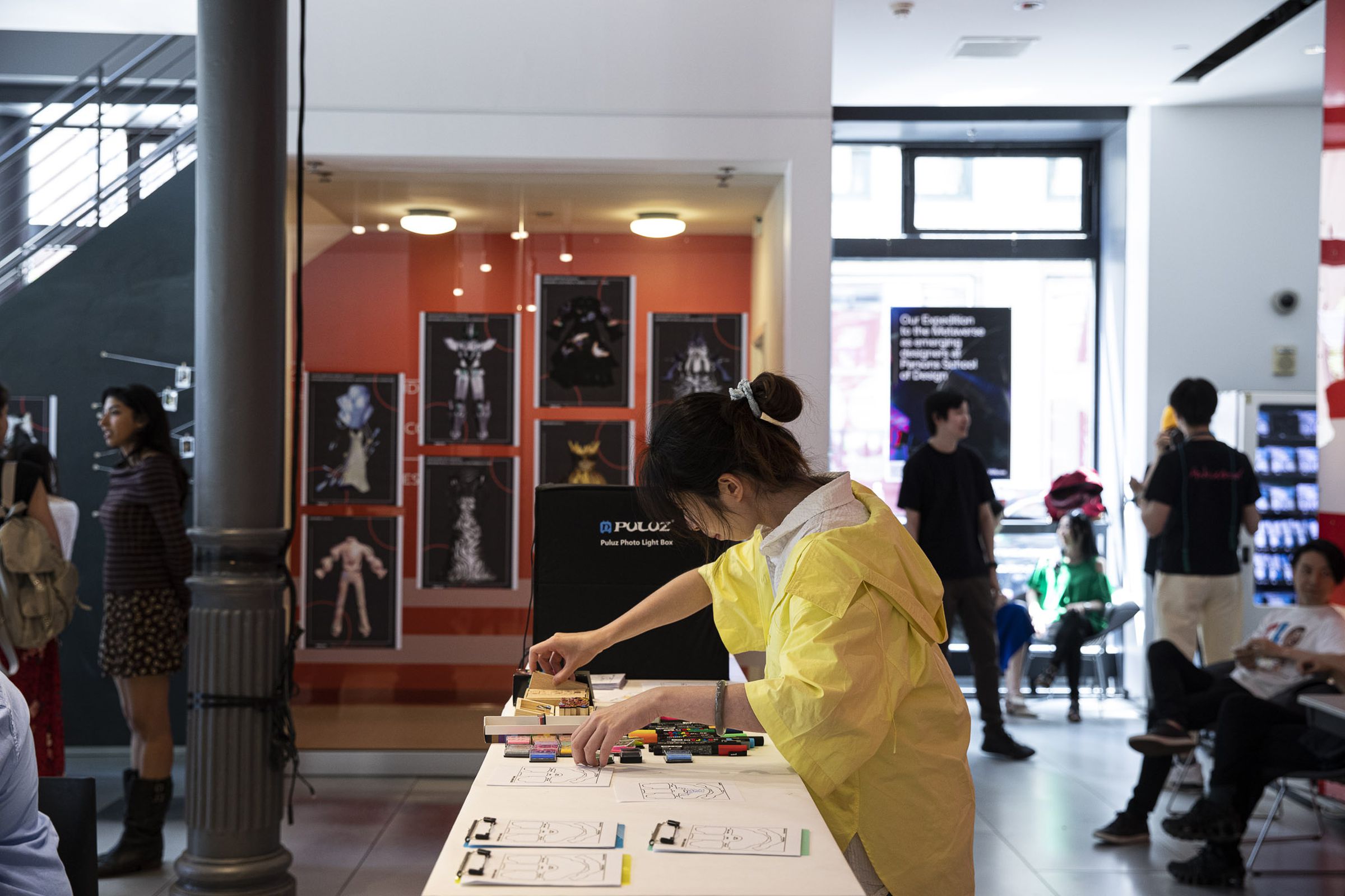 A student works on a drawing project in the foreground. Physical prints of digital clothing hang on the wall behind them.