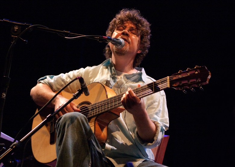 Luciano Antonio plays an acoustic guitar while seated in a chair behind two microphones, one on his guitar and the other for him to sing into