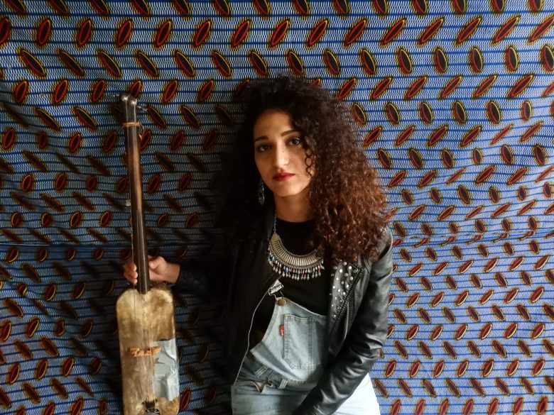 Bab L’ Bluz front woman Yousra Mansour poses against a patterned tapestry holding a guimbri by its neck