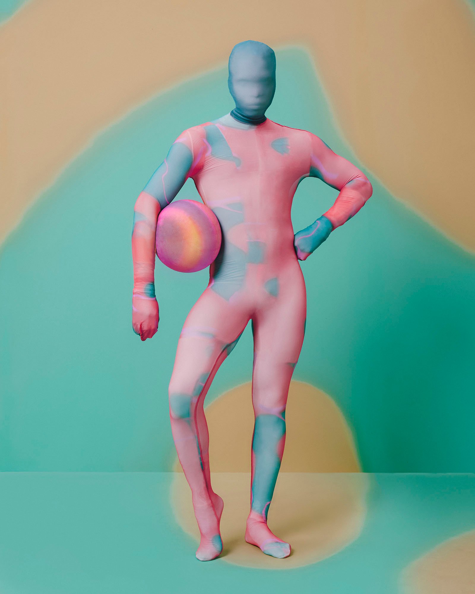 A person covered in a multicolored full-body suit holds a matching ball by their waist.