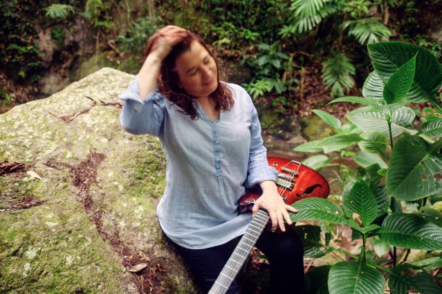 Jana Herzen sitting outside on a mossy rock holding a red bass guitar