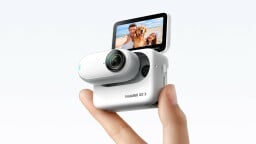 Two fingers holding up the Insta360 GO 3 action camera. It is a white, elongated, thumb-sized oval with a small black lens. The device is inside the Action Pod, a white box with a screen that flips up.