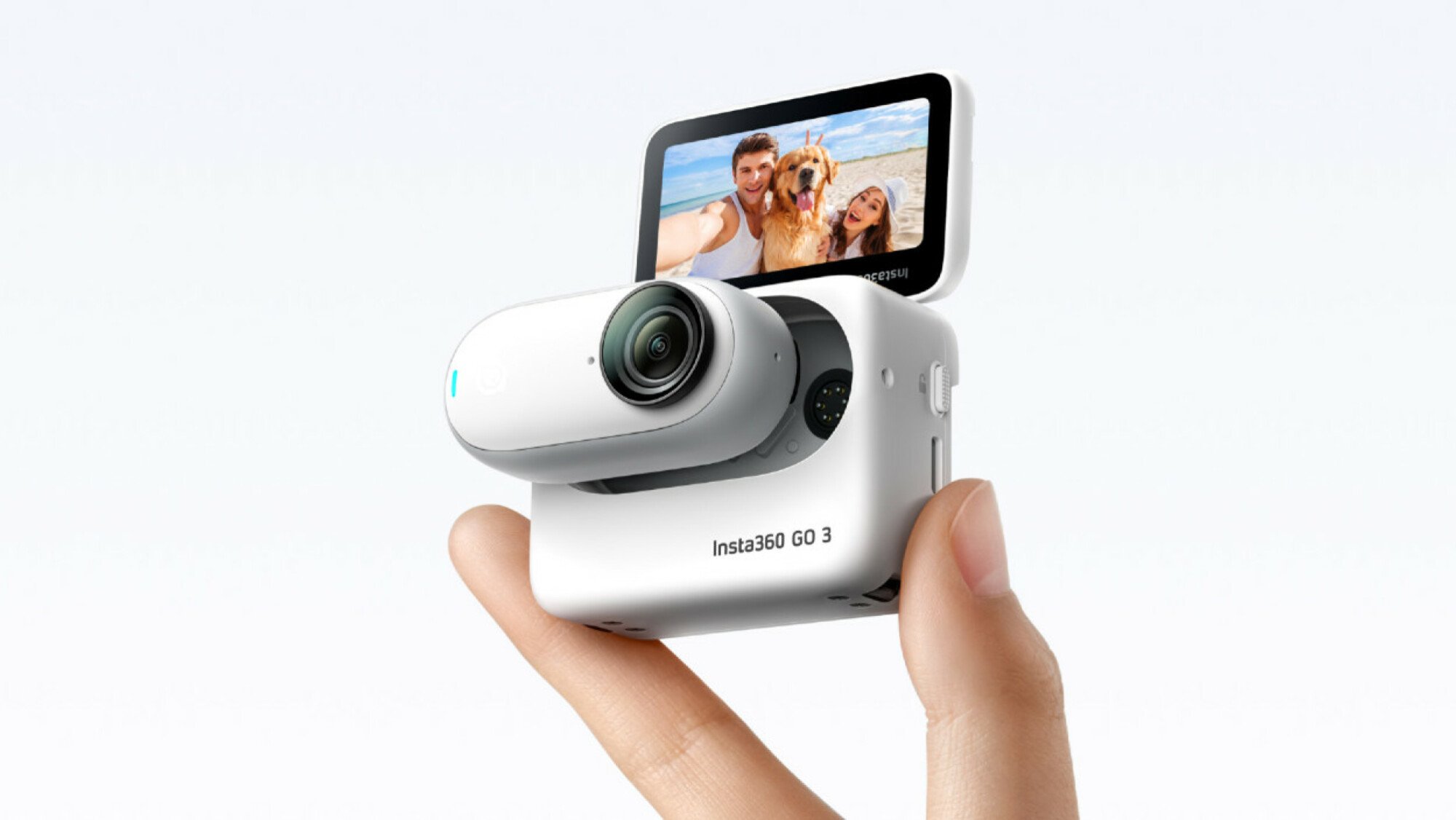 Two fingers holding up the Insta360 GO 3 action camera. It is a white, elongated, thumb-sized oval with a small black lens. The device is inside the Action Pod, a white box with a screen that flips up.