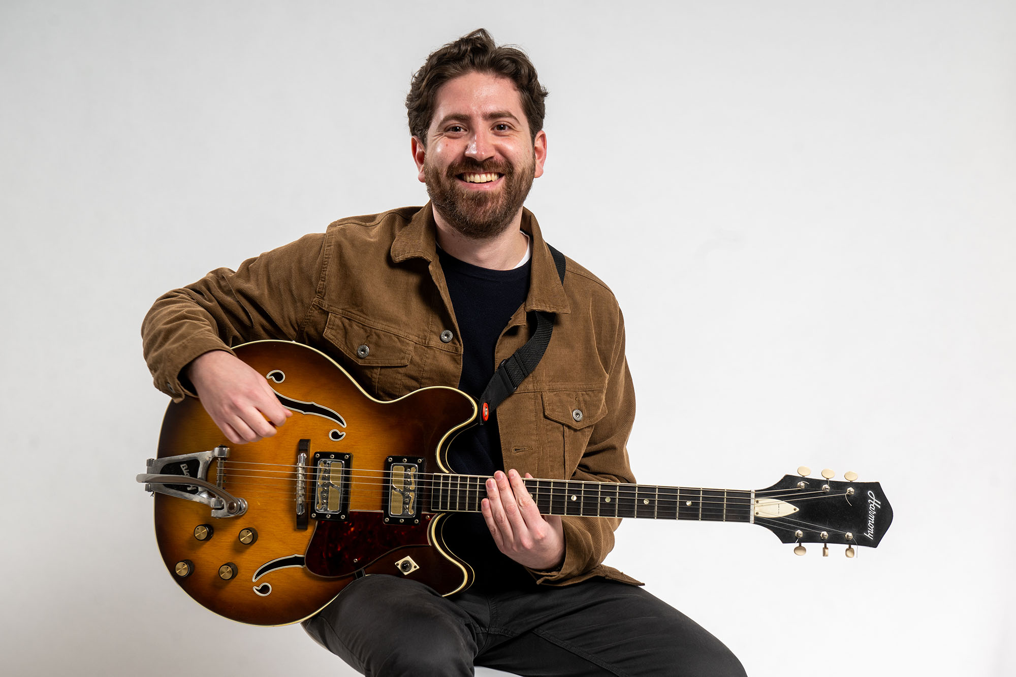 Photo: Erik Broess, a white man white short brown hair and wearing a black shirt, brown jacket, and black pants, smiles and poses with a guitar. He sits in front of a white background.