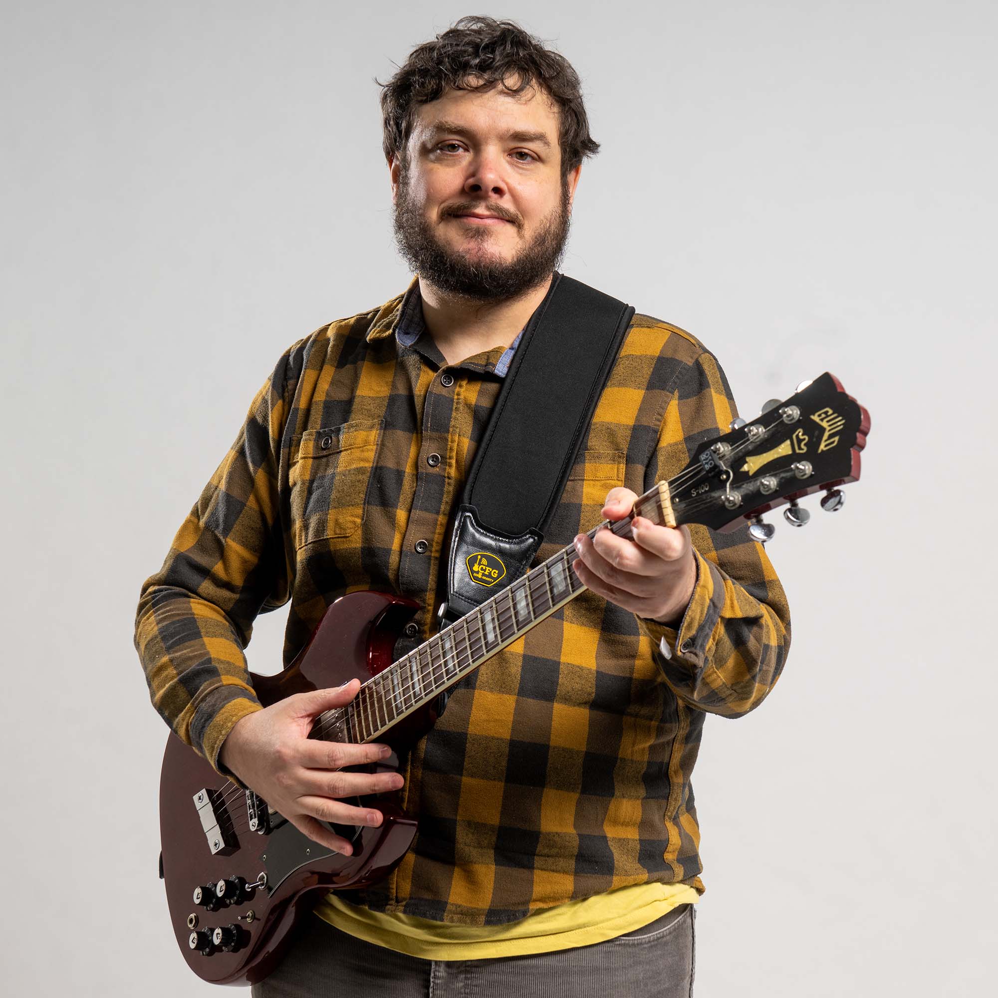 Photo: Lance Morrison, a white man with curly brown hair and wearing a yellow and black plaid collared shirt and grey pants, stands and poses with a guitar in front of a white background.