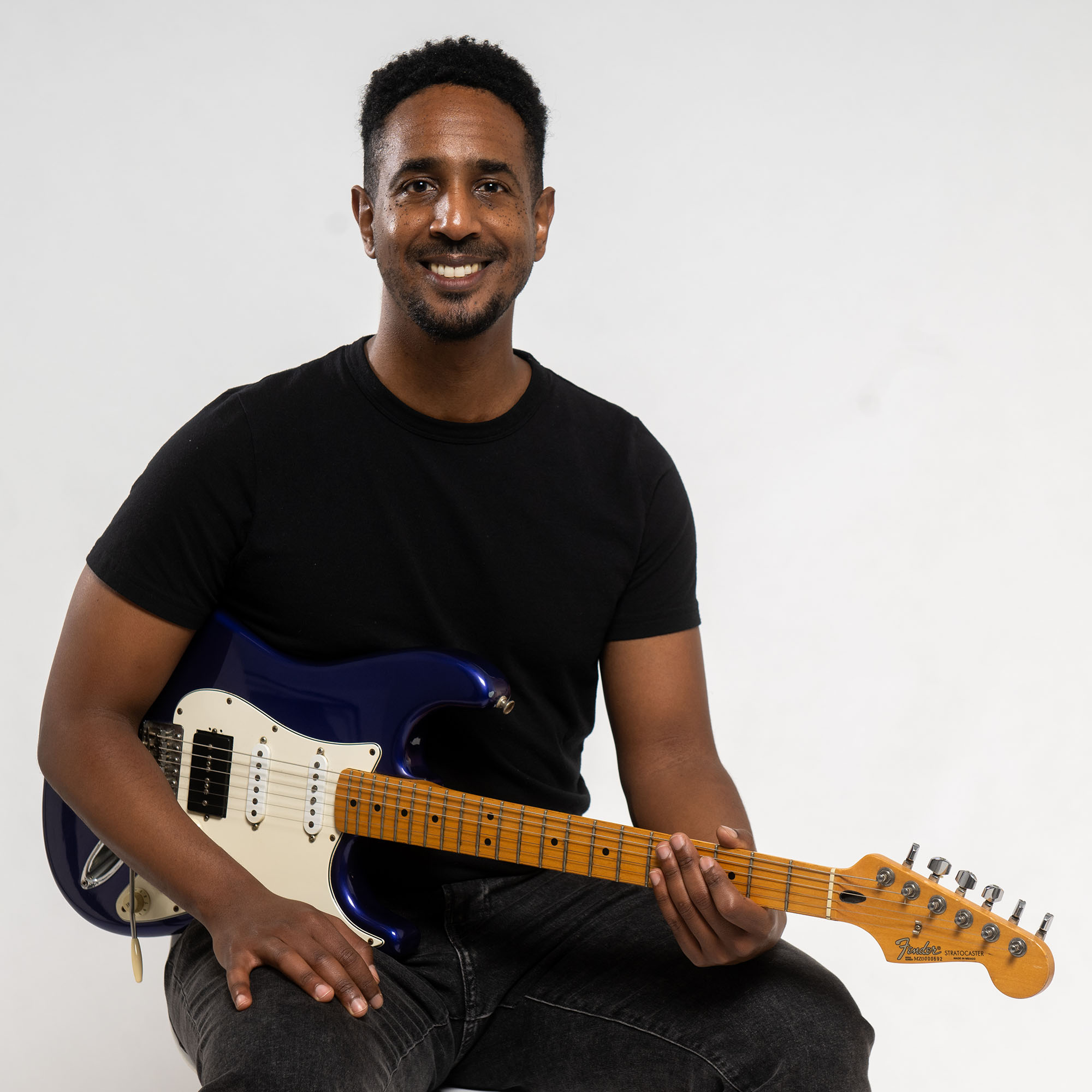 Photo: Kumera Zekarias, a Black man wearing a short-sleeved black shirt and black jeans, smiles and poses with a guitar in front of a white background.