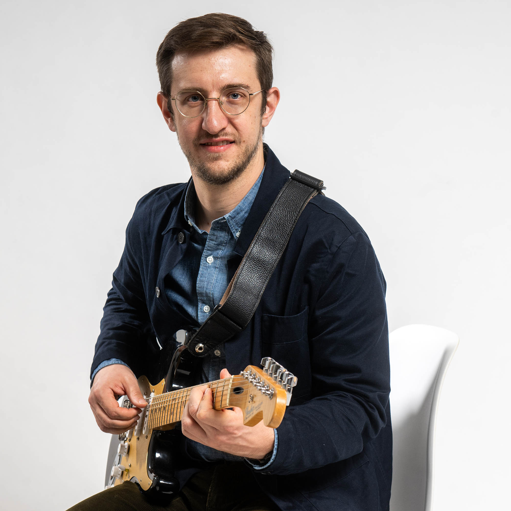 Photo: Brian Barone, a white man with short brown hair and wearing glasses, a blue collared shirt, navy blue jacket, and brown pants, smiles and poses with a guitar. He sits in front of a white background.